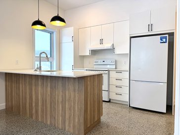 Cuisine moderne et minimaliste avec armoires blanches, poignées noires, cuisinière électrique blanche et réfrigérateur pleine hauteur intégré près de la porte d’entrée.