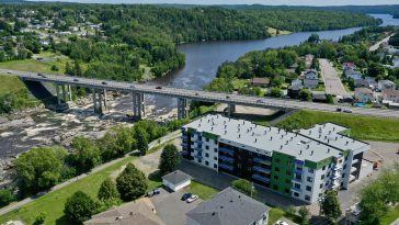Vue aérienne d’un immeuble résidentiel moderne à logements multiples, situé près d’une rivière et d’un pont routier, entouré de verdure et d’un quartier résidentiel.